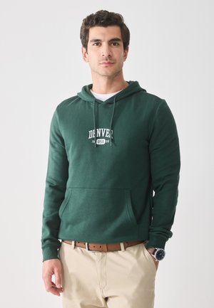 Mann iført en grønn "Denver USA" hettegenser, beige bukser, brun belte og klokke på håndleddet, stående med den ene hånden i lommen på en enkel hvit bakgrunn.