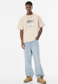 Beige grafisk t-shirt med firkantet print, parret med lyseblå løse jeans og brune snørebåndsstøvler. Detalje med sølv kædetrøje.