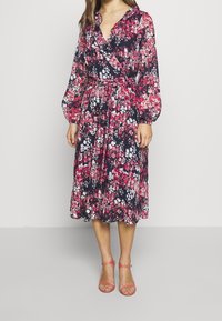 Blommig omslagsklänning i marinblått med rosa och vita mönster. Har en V-ringning, långa ballongärmar och ett knytband i midjan. Lättviktig tyg.