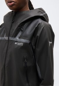 Κοντινό πλάνο ενός ατόμου που φοράει μαύρο αδιάβροχο μπουφάν Columbia Titanium με κουκούλα και φερμουάρ, εμφανίζοντας το λογότυπο και το φερμουάρ της τσέπης στο στήθος.