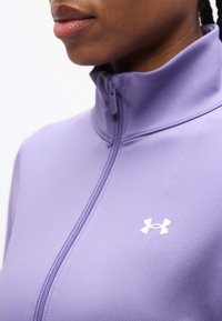 Κοντινό πλάνο ενός προσώπου που φορά ένα μωβ μπουφάν με φερμουάρ και λευκό λογότυπο Under Armour στο στήθος.