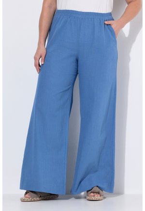 Pantalon large bleu à coupe ample avec ceinture élastique, porté avec des sandales beiges, les mains reposant près des poches, sur fond blanc uni.