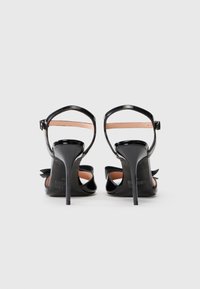 Black high-heeled sandals with a shiny patent finish, ankle strap, and bow accent at the back, featuring a slender stiletto heel.