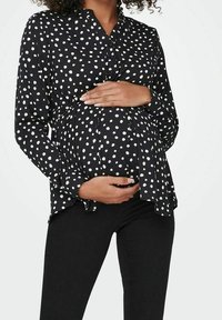 Blusa in maternità a pois neri con bottoni sul davanti, design leggermente svasato e maniche lunghe, abbinata a pantaloni neri aderenti.