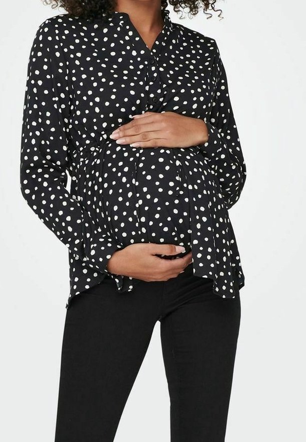 Blusa in maternità a pois neri con bottoni sul davanti, design leggermente svasato e maniche lunghe, abbinata a pantaloni neri aderenti.