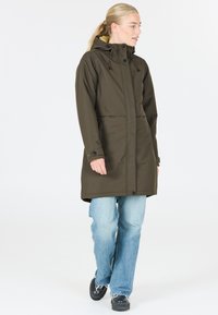 Olijfgroene, knielange jas met capuchon, beschikt over een ritssluiting, drukknoopjes en twee frontzakken. Draagt over lichtblauwe jeans.