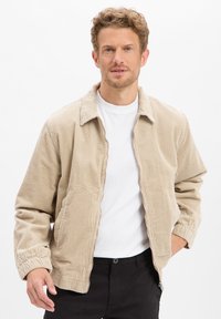 Beige corduroy jack met een ritssluiting aan de voorkant, lange mouwen en een overhemdkraag, gedragen over een wit t-shirt, gecombineerd met zwarte broek.