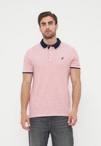 Άνδρας που φοράει ροζ πόλο με μπλε σκούρο γιακά και περίγραμμα στα μανίκια, στέκεται σε απλό λευκό φόντο, κοιτάζοντας μπροστά.