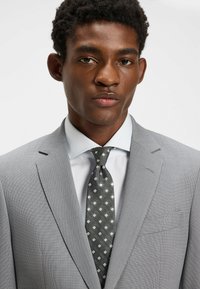 Habillé en costume texturé gris, chemise bleu clair et cravate gris foncé à motifs de diamants blancs. Gros plan sur le haut du corps, mettant l'accent sur les détails des vêtements.