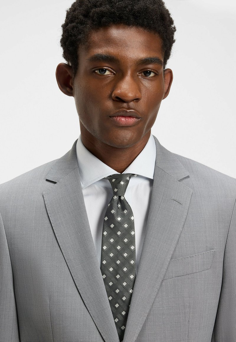 Habillé en costume texturé gris, chemise bleu clair et cravate gris foncé à motifs de diamants blancs. Gros plan sur le haut du corps, mettant l'accent sur les détails des vêtements.