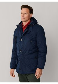 Parka aislada azul marino con capucha, con botones de palanca, bolsillos laterales y textura suave. Camisa roja debajo que añade contraste.