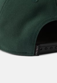 Πράσινο ύφασμα καπέλο με υφή, διαθέτει μαύρη πλαστική ρυθμιζόμενη λουρί και μια μικρή ετικέτα με το "JORDAN" σε λευκά γράμματα.