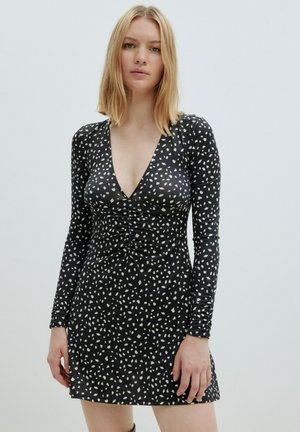 Schwarzes Kleid mit langen Ärmeln, V-Ausschnitt, gerafftem Vorderteil und einem verstreuten weißen Muster. Weicher, enganliegender Stoff mit einem ausgestellten Saum.