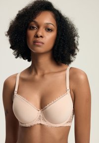 Młoda kobieta z kręconymi włosami, ubrana w biustonosz w kolorze nude z koronkowym wykończeniem, stojąca na tle jasnym.