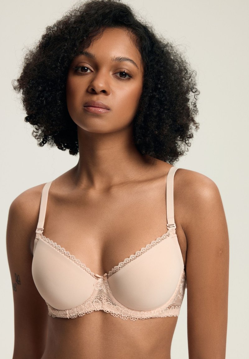 Młoda kobieta z kręconymi włosami, ubrana w biustonosz w kolorze nude z koronkowym wykończeniem, stojąca na tle jasnym.
