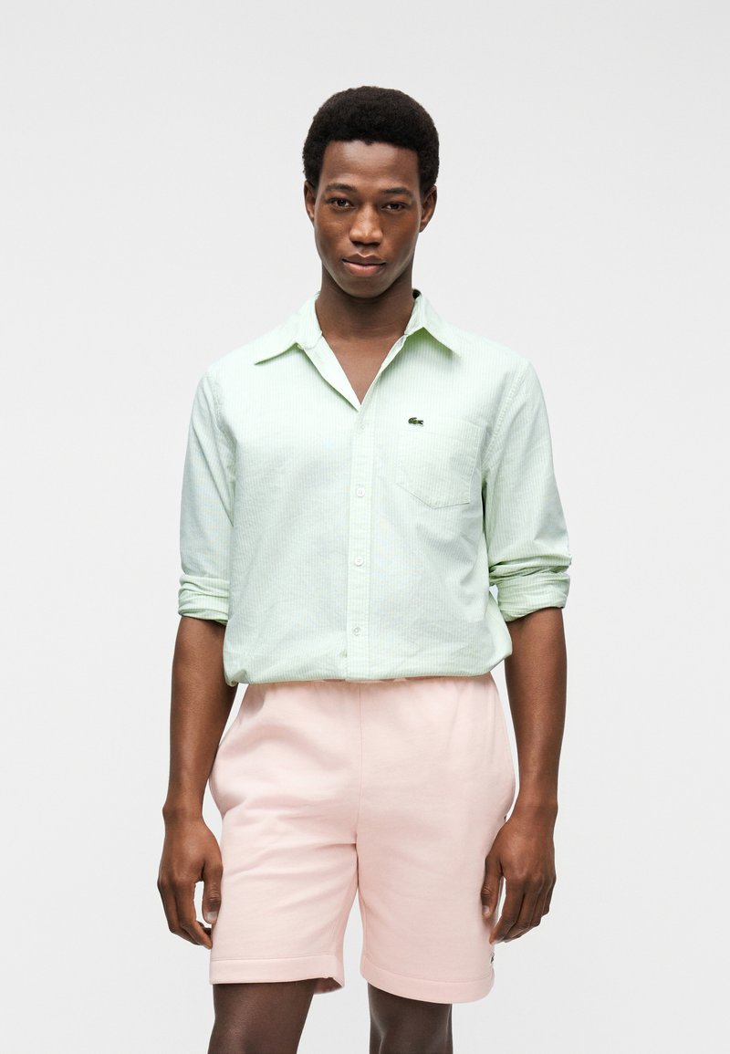 Homme portant une chemise à boutons vert clair aux manches retroussées et un short rose pâle, debout devant un fond blanc.