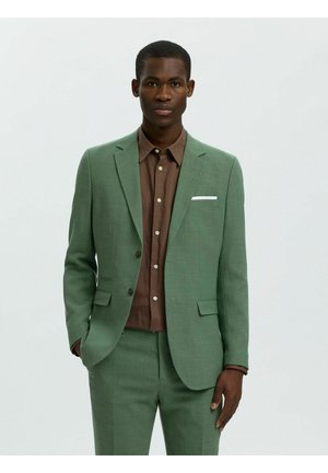 Άνδρας που φοράει πράσινο κοστούμι με λευκή μαντηλάκι στην τσέπη και καφέ πουκάμισο με κουμπιά, στέκεται με το ένα χέρι στην τσέπη.