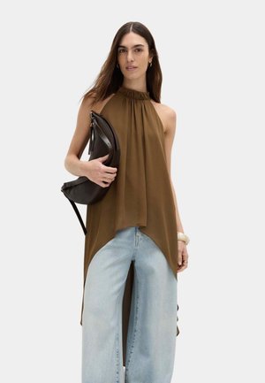 Mujer vestida con una túnica marrón sin mangas de cuello alto y jeans anchos de pierna azul claro, sosteniendo un bolso negro sobre un fondo liso.