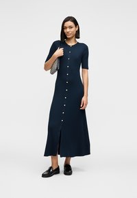 Ναυτικό μπλε ριγέ φόρεμα με κουμπιά μπροστά, κοντά μανίκια, στρογγυλή λαιμόκοψη, πλευρική σχισμή και κουμπιά σε χρυσαφί απόχρωση. Συνδυάζεται με μαύρα παπούτσια και ασημένια τσάντα.