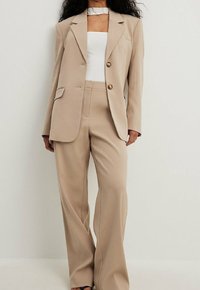 Blazer beige su misura con due bottoni, rever a taglio e tasche laterali, abbinato a pantaloni a gamba larga nello stesso tessuto liscio.