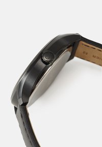 Reloj de acero inoxidable negro con un acabado suave, caja redonda y una corona texturizada. Correa de cuero negro con detalles de costura clara.