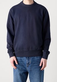 Tmavě modrá mikina s kulatým výstřihem, žebrovanými manžetami a lemem. Má jemné značkování na límci a měkký materiál z bavlněné směsi.