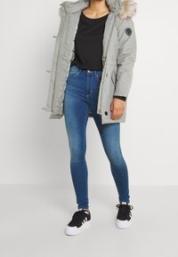 Parka gris con capucha forrada de piel, camiseta negra, jeans ajustados azules y zapatillas negras. Presenta una cremallera y múltiples bolsillos.