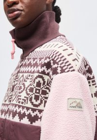 Une personne porte un pull à motifs avec des motifs géométriques violets et blancs, un col montant avec un cordon rose et un patch en polaire rose sur la manche avec un logo.