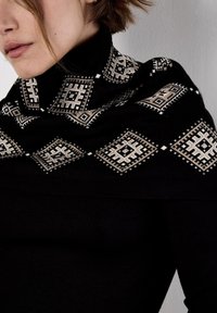 Dolcevita nero con scialle decorato che presenta disegni geometrici beige e sottili decorazioni lungo i bordi. Texture in morbido tessuto a maglia.