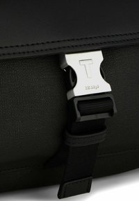 Borsa nera in tessuto con superficie testurizzata, dotata di una fibbia in metallo argentato con il logo "T" e un dettaglio di cinturino cucito.