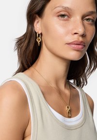 Guldøreringe i hoop-design med sammenkoblede elementer, parret med en guldkædehalssmykke med en dobbeltloop pendants, båret med en beige ribbet top.