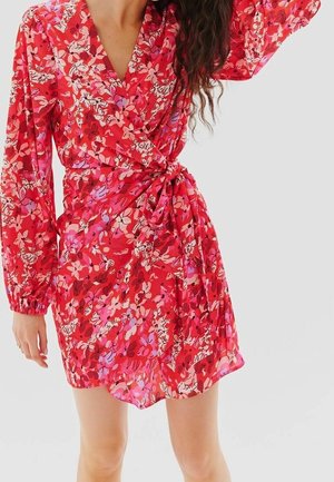 Kobieta w czerwonej sukience kopertowej w kwiaty z długimi bufkami, przewiązanej w talii, stojąca na jasnym, jednolitym tle.