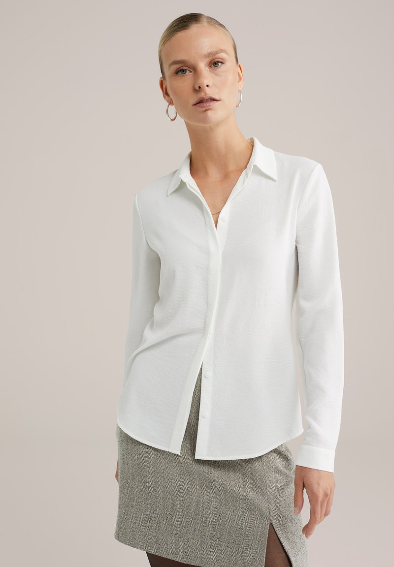 Donna che indossa una camicia bianca a maniche lunghe con bottoni, una gonna grigia strutturata con spacco frontale e orecchini a cerchio, su uno sfondo neutro.