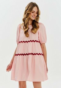 Różowa sukienka z krótkim rękawem i okrągłym dekoltem, z falbaniastymi warstwami i kontrastowym bordowym wykończeniem w zygzak na wysokości talii. Gładka faktura tkaniny.