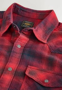 Rød og sort ternet flannel skjorte med en button-down krave. Har metaltrykknapper, et vævet mærke og et blødt, tekstureret stof.