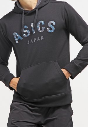 Czarna bluza z kapturem z przednią kieszenią, niebieskim kamuflażowym logo ASICS i szarym napisem Japan, noszona z czarnymi spodniami.