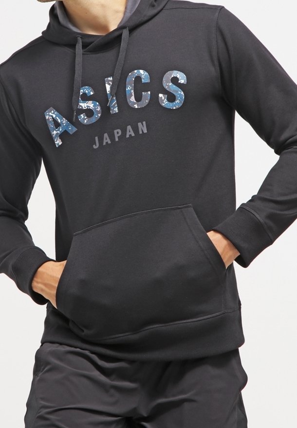 Sweat à capuche noir avec poche frontale, logo ASICS camouflage bleu et texte gris « Japan », porté avec un pantalon noir.