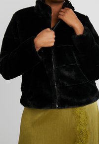 Chaqueta de piel sintética negra con cremallera y cuello alto, que presenta costuras horizontales y una textura suave. Llevada sobre una falda verde de canalé con detalles de encaje.