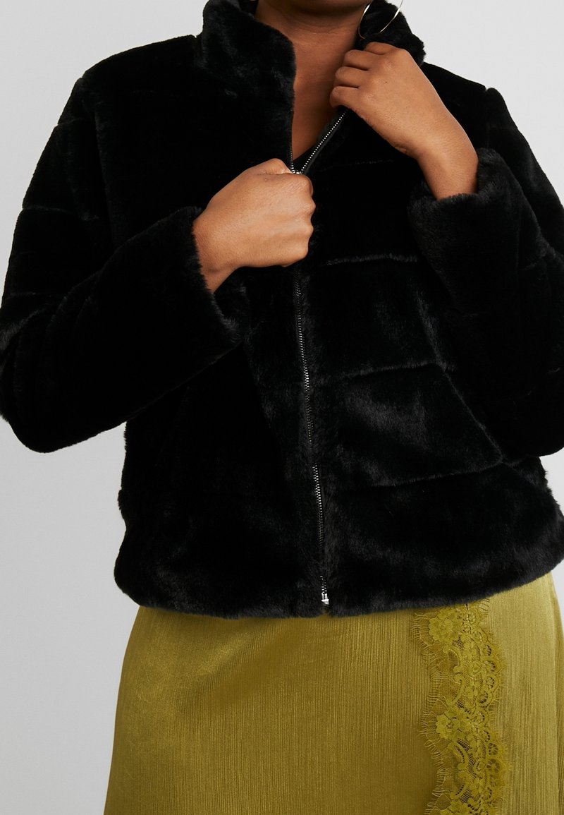 Chaqueta de piel sintética negra con cremallera y cuello alto, que presenta costuras horizontales y una textura suave. Llevada sobre una falda verde de canalé con detalles de encaje.