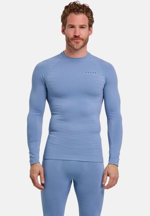 Uomo con capelli ricci che indossa una maglia aderente a maniche lunghe azzurro chiaro e leggings abbinati, in piedi davanti a uno sfondo neutro.