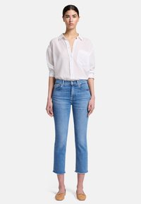 Camicia bianca leggera con taschino sul petto, abbinata a jeans azzurri a vita alta, tagliati alla caviglia. Scarpe flat marroni.