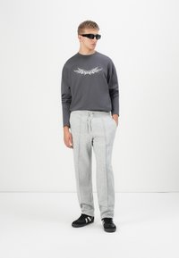 Sudadera gris de cuello redondo con gráfico tribal, combinada con pantalones de chándal gris claro que tienen cordón y ribetes laterales, llevados con zapatillas negras.