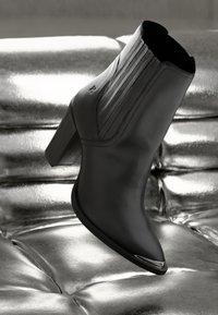 Melnas ādas potīšu zābaki ar augstu papēdi, ar vertikālām šuvēm un spīdīgu sudraba akcentu uz purngala, uz metāliski spīdīga fona.