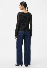 Maglietta nera a maniche lunghe con ricami floreali, in tessuto trasparente, abbinata a jeans blu a vita alta e a gamba ampia, con scarpe nere a punta.