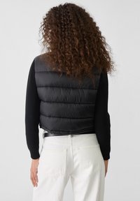 Doudoune noire sans manches avec des coutures horizontales, associée à un pull côtelé noir. Le mannequin porte un jean taille haute blanc avec des poches arrière.