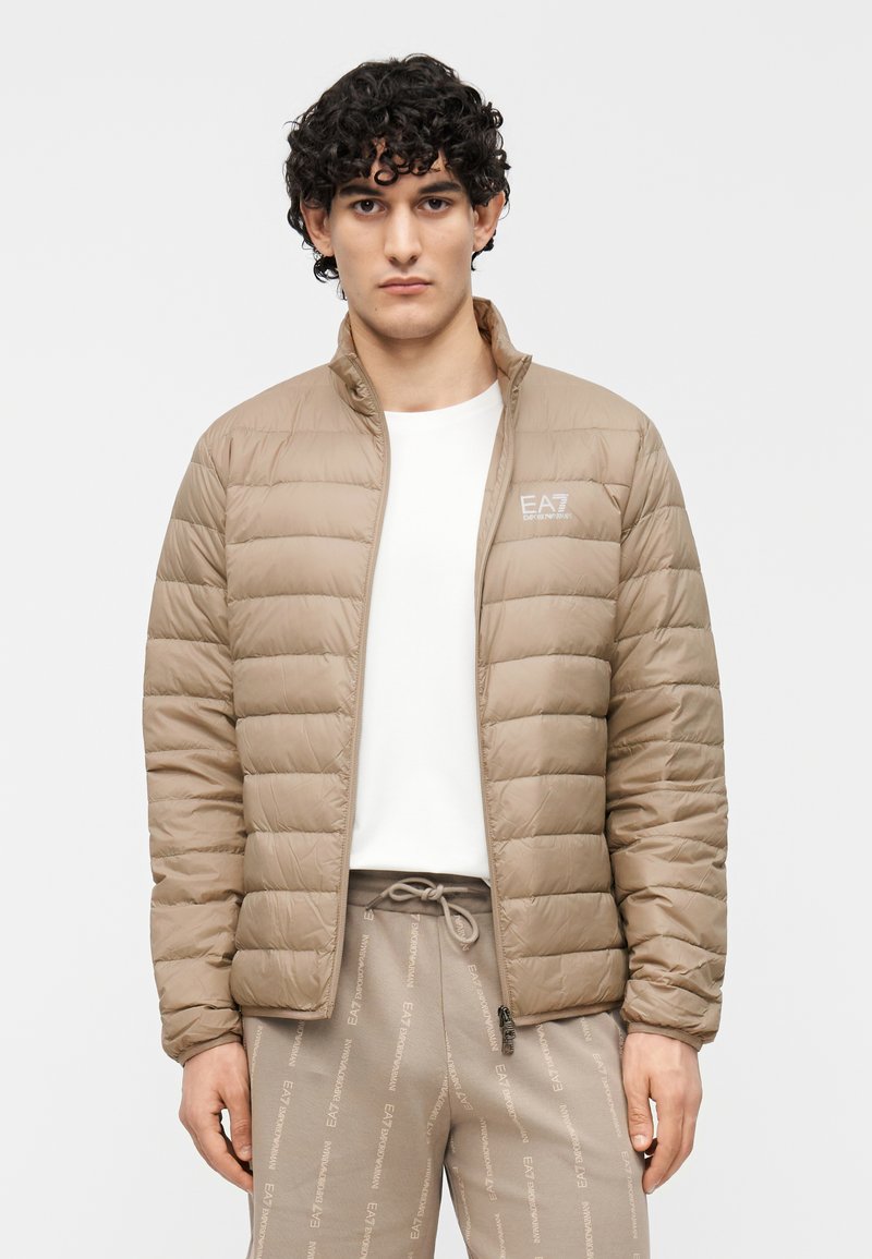 Bézs színű steppelt puffertalp kabát cipzáras elülső résszel, magas gallérral és vízszintes steppeléssel. Könnyű anyag, világos színű nadrággal párosítva.