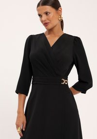 Czarna sukienka kopertowa z rękawami o długości trzech kwartałów, fakturowana tkanina, talia podkreślona, oraz złoty okrągły detal z klamrą. Gładka, elegancka sylwetka.