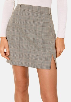 Beige-schwarzer karierten Minirock mit kleinem Frontschlitz, getragen mit einem leichten langärmeligen Oberteil, das bis zur Mitte des Oberschenkels reicht und Hände liegen in der Nähe.