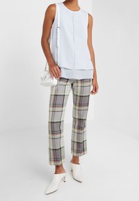 Kvinna klädd i en ärmlös ljusblå topp, grå rutiga byxor med gula och rosa ränder, vita klackskor och en vit axelväska.