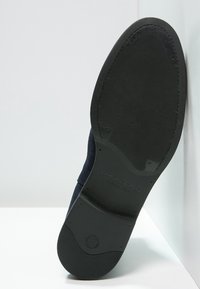 Suela de goma negra de un zapato azul oscuro, con una superficie texturizada y una marca en la entresuela. Vista trasera sobre un fondo claro.